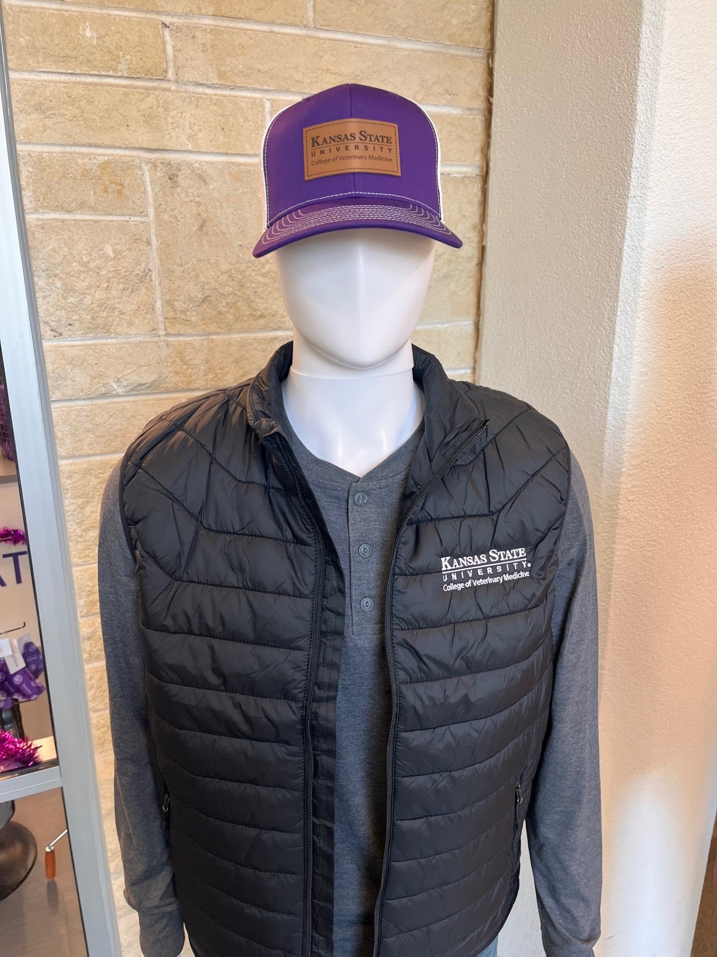 Mannequin wearing a black puffer vest and gray shirt with a purple and white trucker hat with Kansas State University College of Veterinary Medicine logo patch.