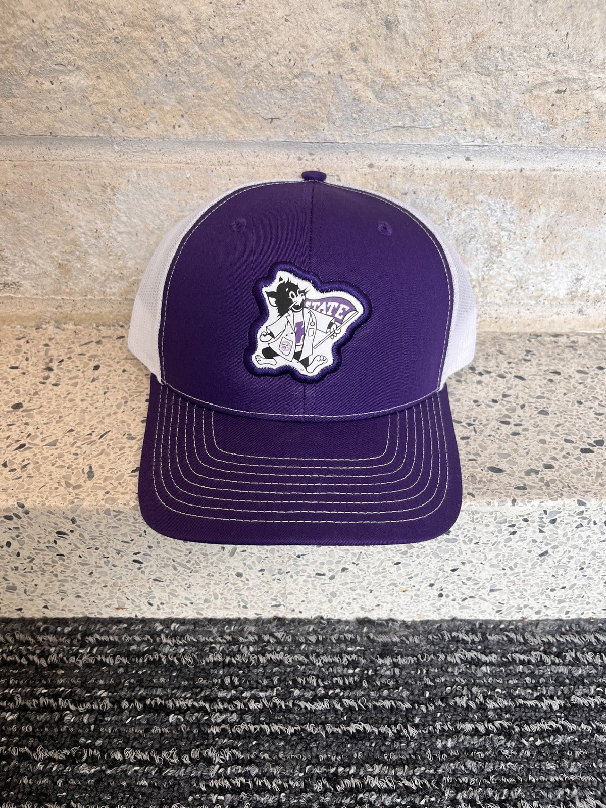A purple and white trucker hat with a custom embroidered patch on the front, displayed on a speckled stone ledge.