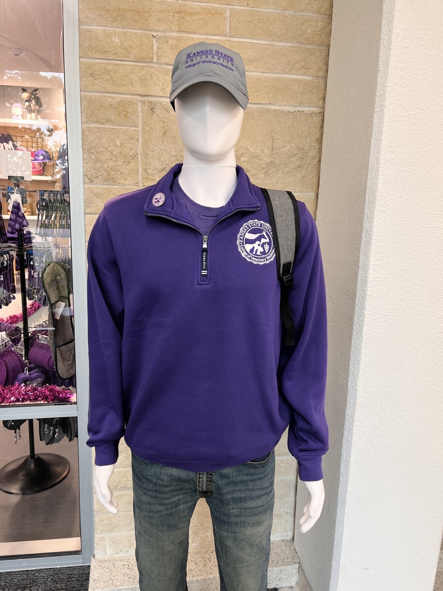 A mannequin wearing a purple quarter-zip sweatshirt with a circular white logo on the left chest, paired with gray jeans and a gray baseball cap. The mannequin is positioned in front of a beige stone wall and a store window.