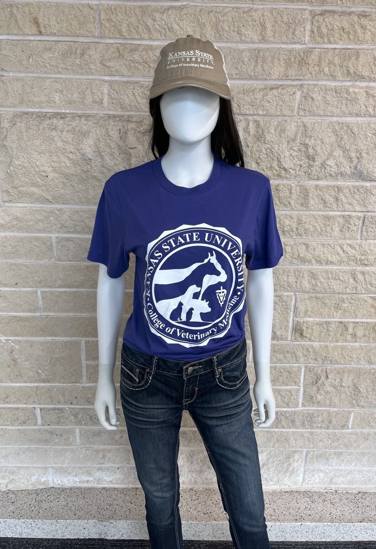 A mannequin wearing a purple t-shirt with the College of Veterinary Medicine Seal Logo, paired with dark wash jeans and a tan baseball cap, set against a beige brick wall.