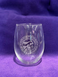 A clear stemless wine glass with an etched CVM seal in the center, against a purple background. 