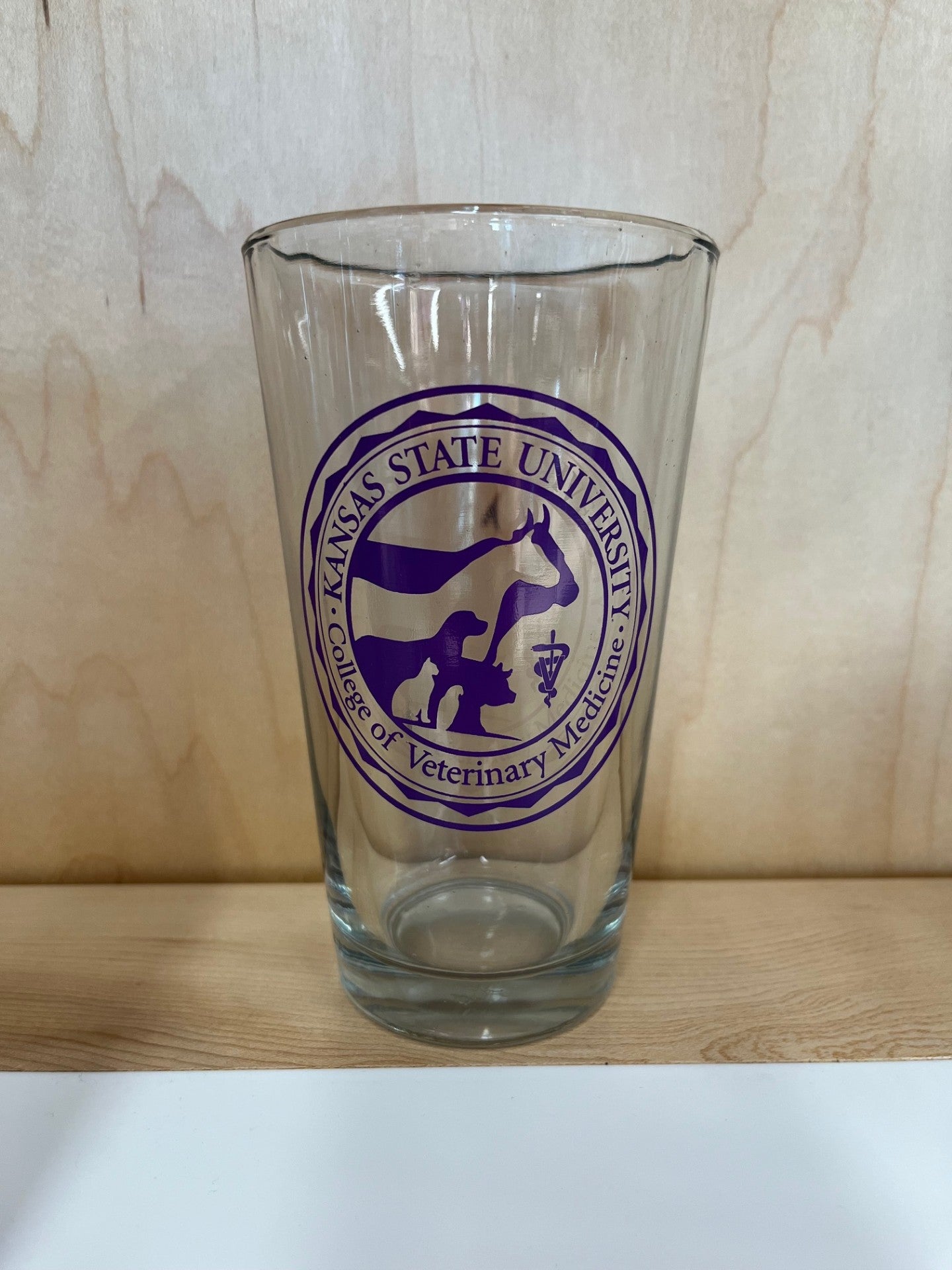 A clear pint glass with a purple Kansas State University College of Veterinary Medicine logo in center.  