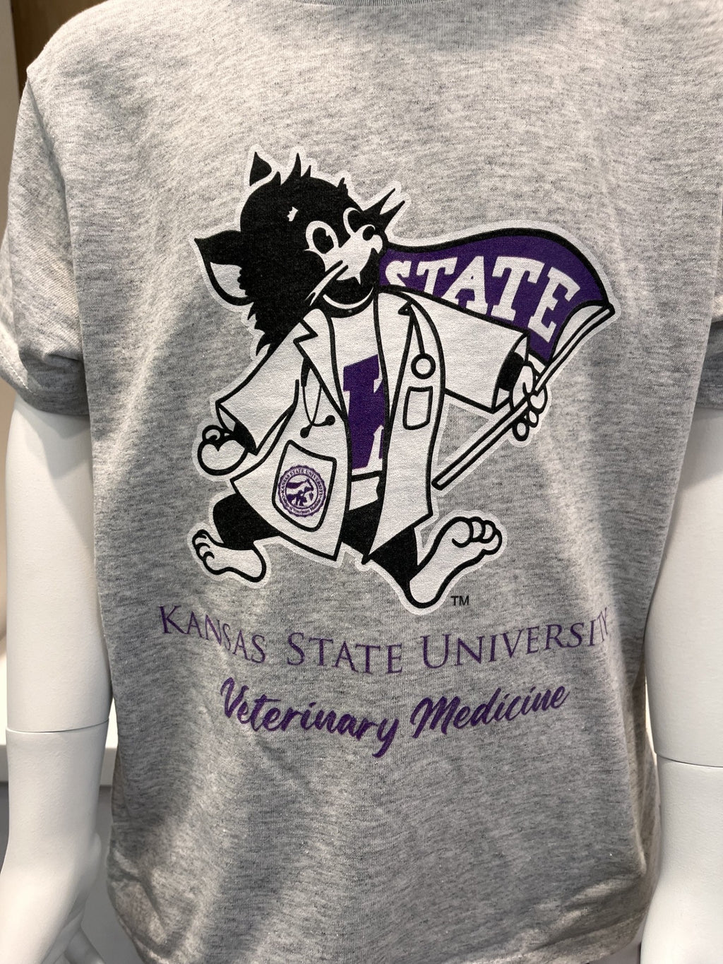 A heather gray short-sleeve t-shirt displayed on a white mannequin torso, featuring a large graphic of Lab Coat Willie Below the logo, the text 'KANSAS STATE UNIVERSITY' and 'Veterinary Medicine' is printed in purple.