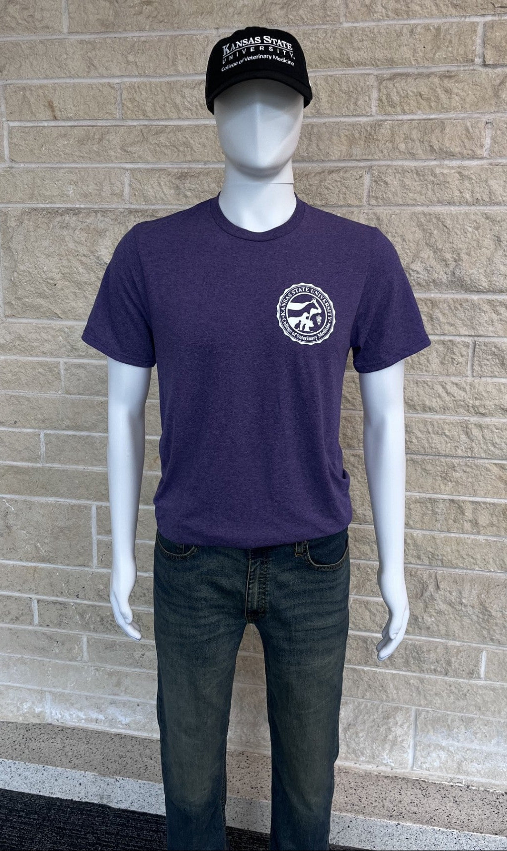 A mannequin wearing a dark purple heathered t-shirt with a white circular graphic on the left chest, paired with dark gray jeans and a black baseball cap, set against a beige brick wall background.