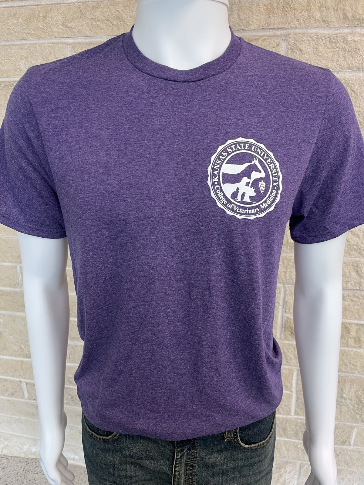 A heather purple short-sleeve t-shirt displayed on a white mannequin torso, featuring a white circular logo printed on the left chest, paired with dark blue jeans against a light brick wall background.