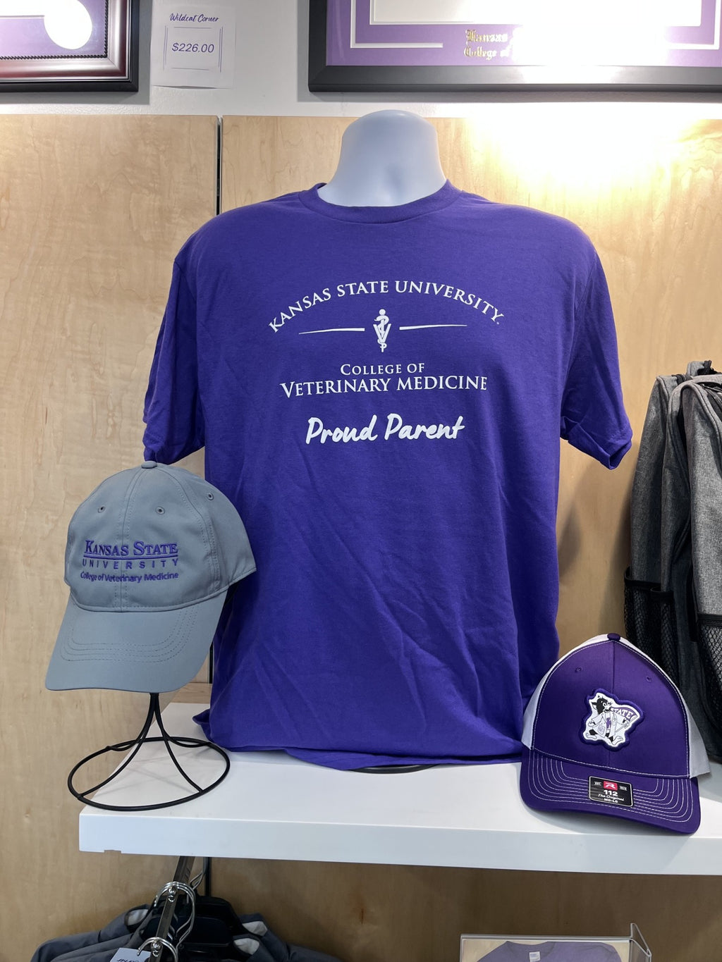 A purple short-sleeve T-shirt displayed on a mannequin torso in a retail setting, printed with white text reading 'Kansas State University College of Veterinary Medicine Proud Parent.' Two matching baseball caps are also shown on stands: one light gray and one purple, both with Kansas State branding.