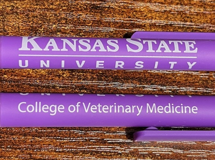 Two purple pens with 'Kansas State University College of Veterinary Medicine' text on a wooden surface