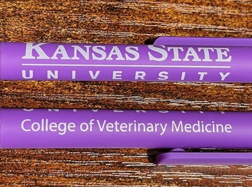 Two purple pens with 'Kansas State University College of Veterinary Medicine' text on a wooden surface