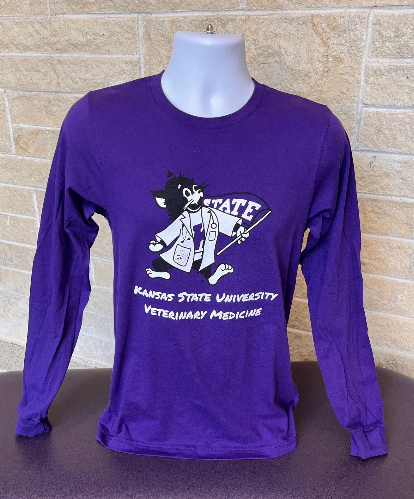Purple long-sleeve shirt with a Labcoat Willie graphic and "Kansas State University Veterinary Medicine" text on a mannequin against a brick wall.