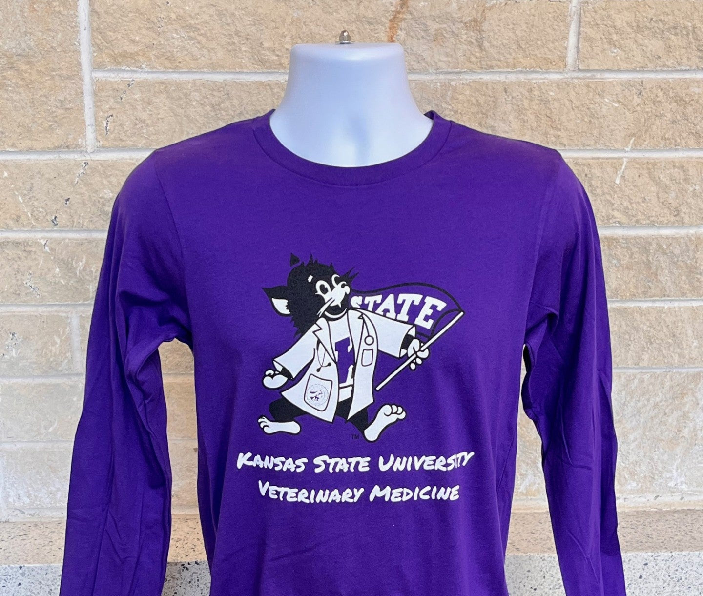 Purple long-sleeve shirt with a Labcoat Willie graphic and "Kansas State University Veterinary Medicine" text on a mannequin against a brick wall.