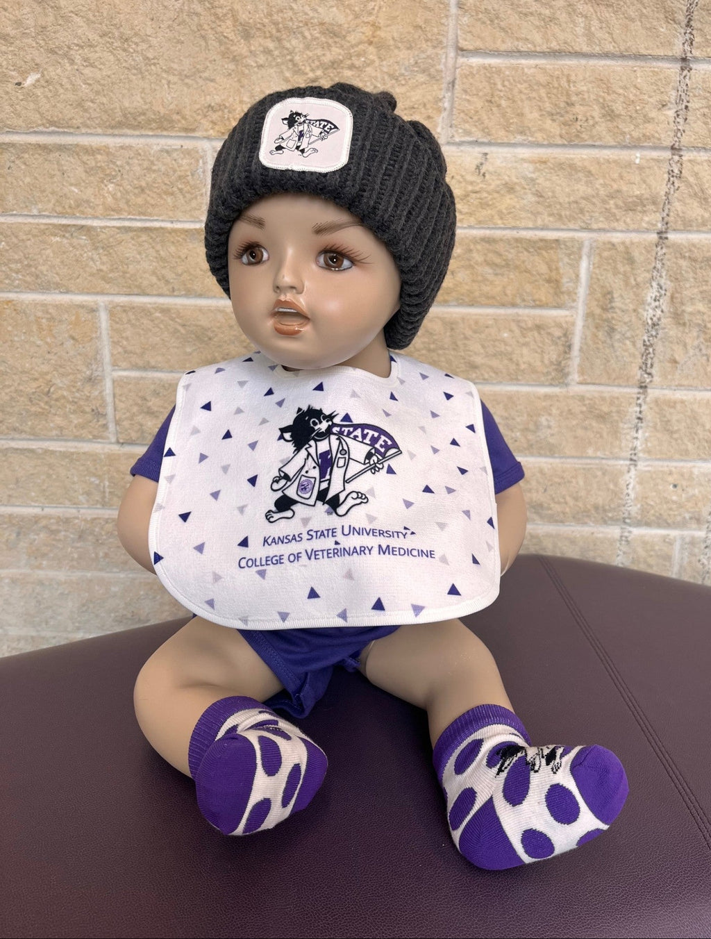 Baby doll wearing a Kansas State University College of Veterinary Medicine bib and purple socks against a brick wall.