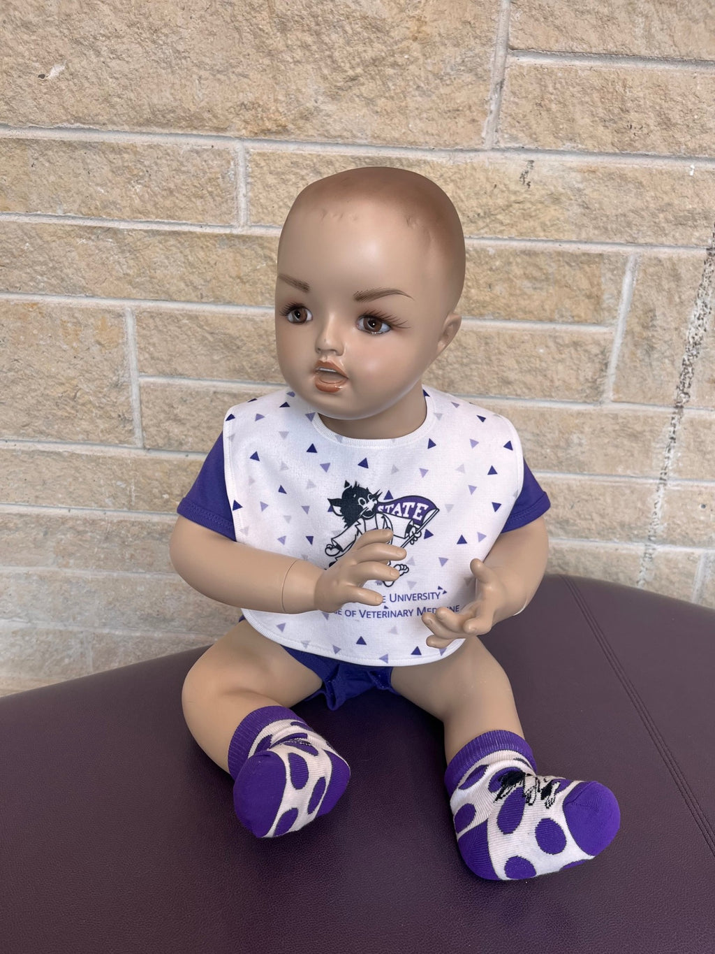Baby doll wearing a purple outfit with a bib, sitting on a table against a brick wall.