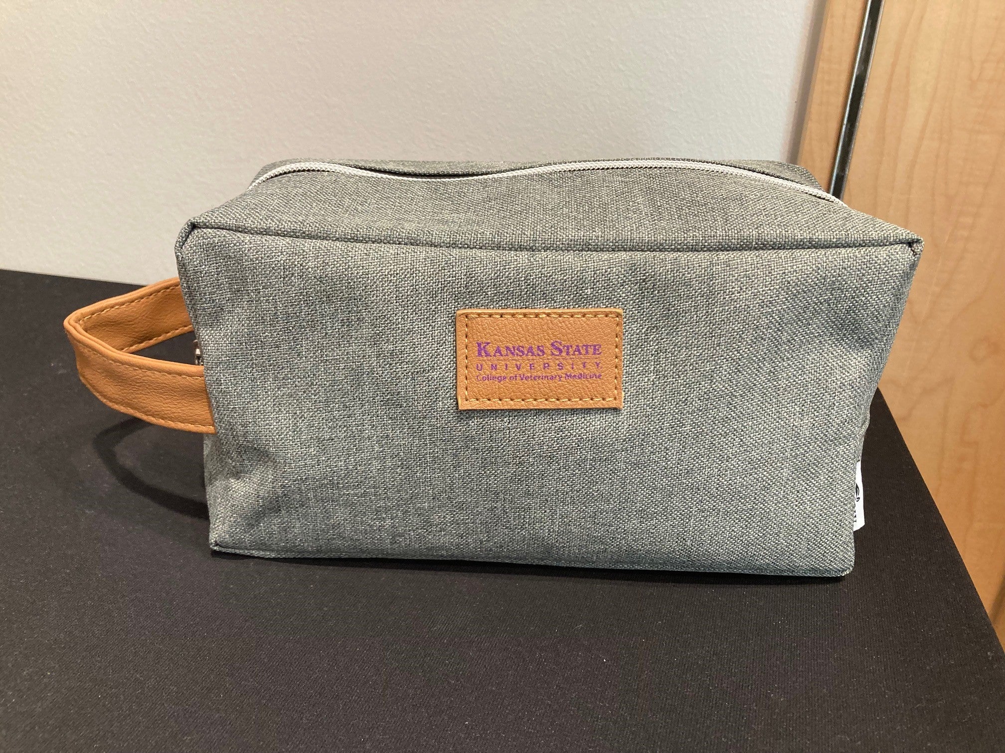 A gray toiletry bag with a brown leather handle and a brand leather label that reads 'Kansas State University College of Veterinary Medicine'.