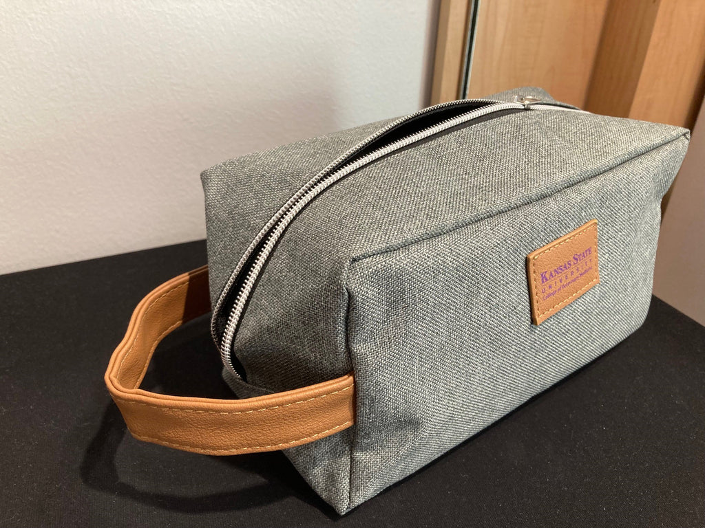 A gray toiletry bag with a brown leather handle and a brand leather label that reads 'Kansas State University College of Veterinary Medicine'.
