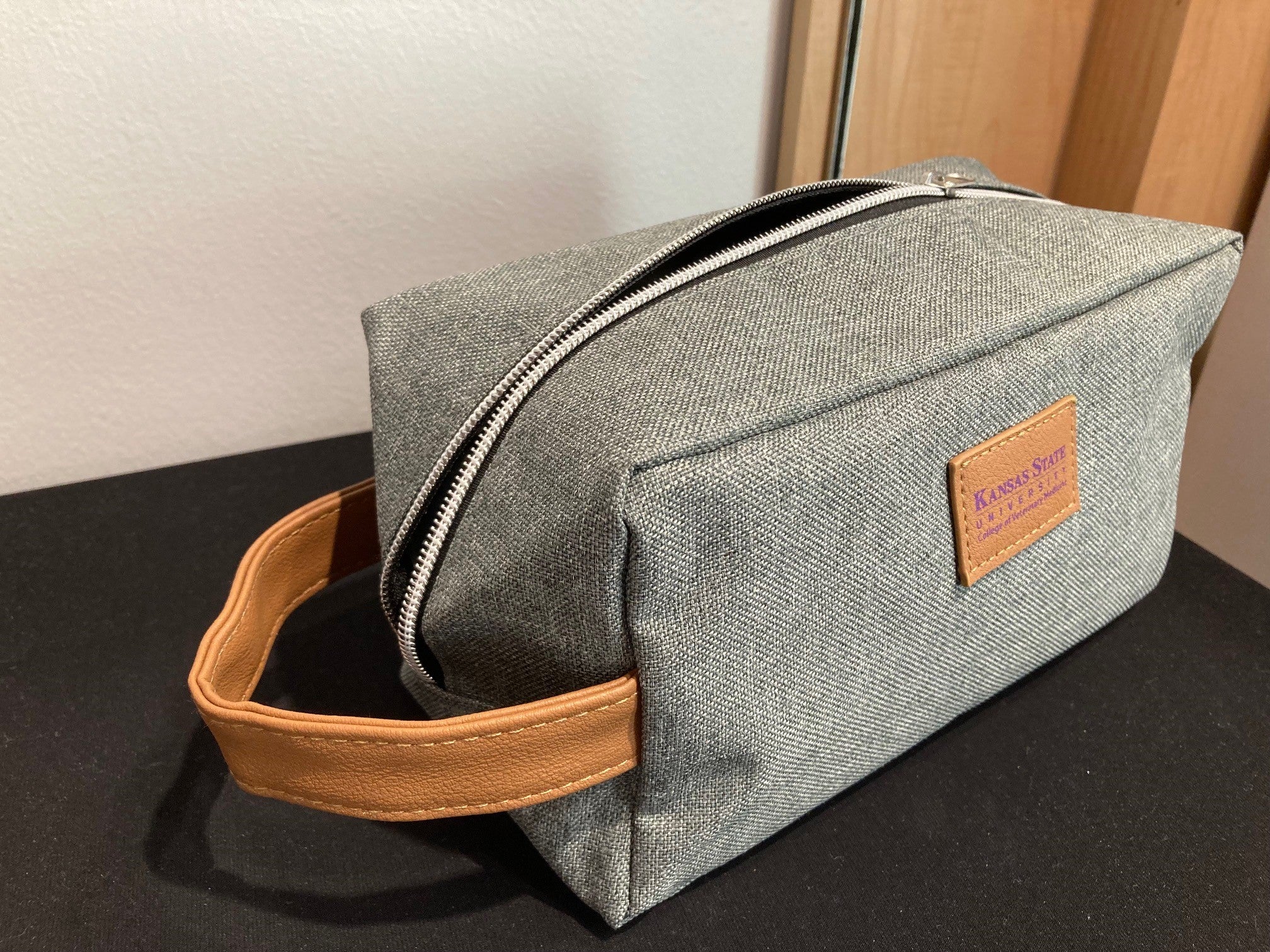 A gray toiletry bag with a brown leather handle and a brand leather label that reads 'Kansas State University College of Veterinary Medicine'.