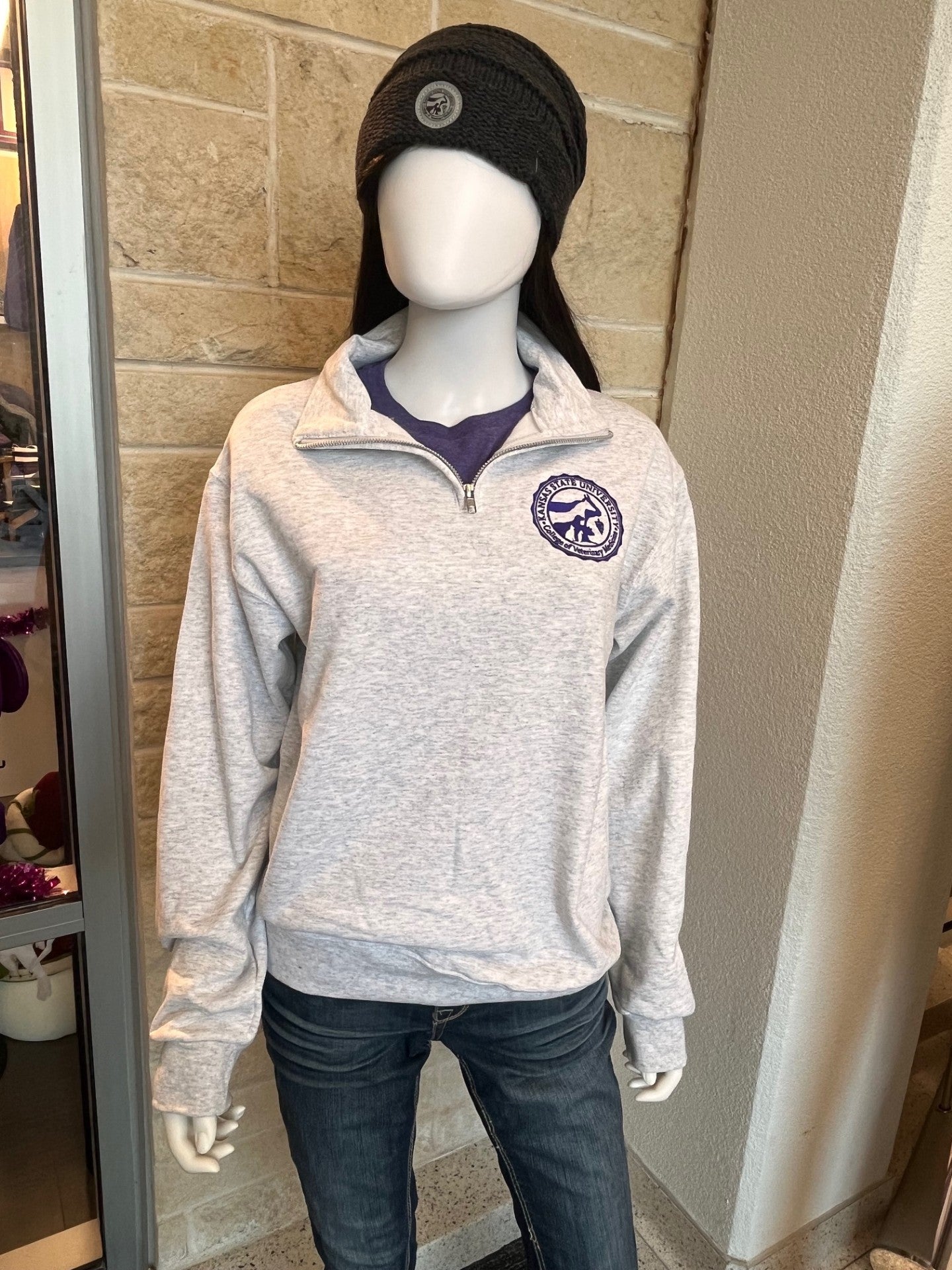 Mannequin wearing a gray pullover with a purple CVM seal on the left chest, black beanie, and dark jeans against a stone wall.
