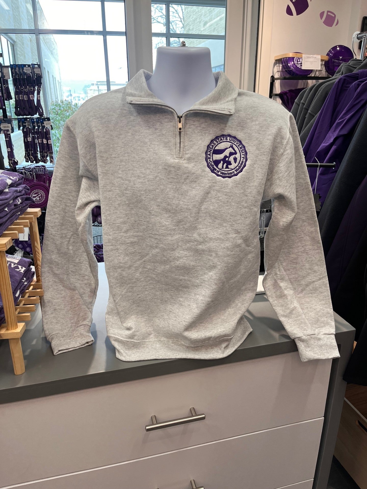 Mannequin wearing a gray pullover with a purple CVM seal on the left chest in a store setting. 