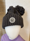 A chunky knit beanie with a pom-pom on a mannequin head, featuring a leather CVM seal patch on the front.