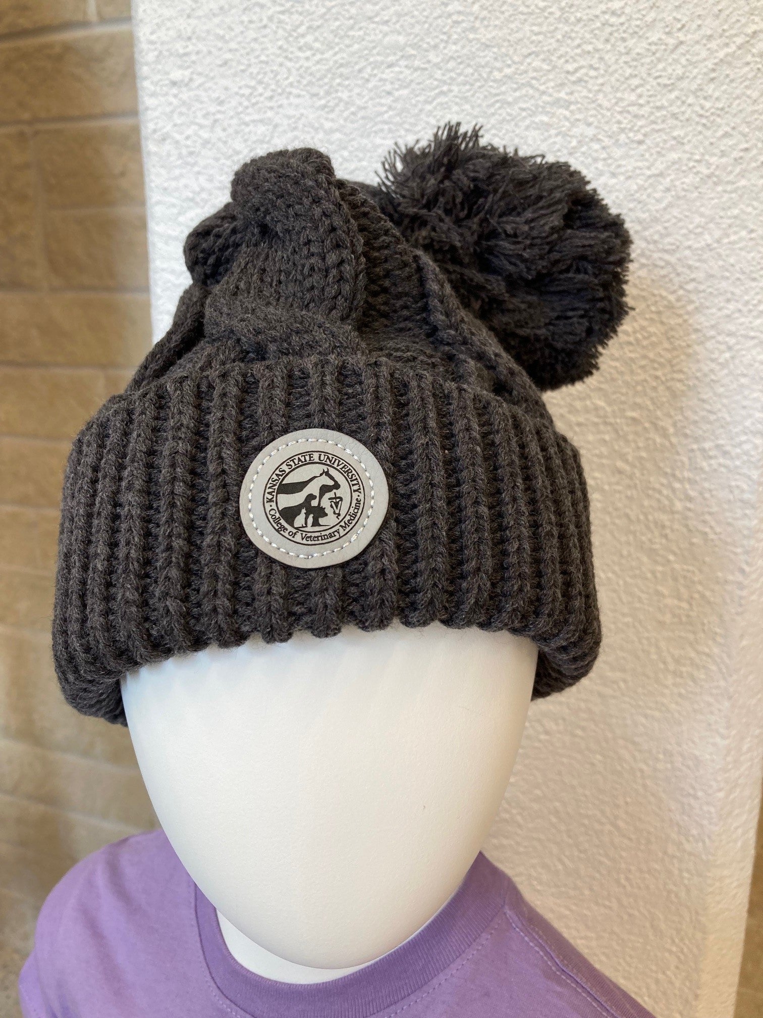 A chunky knit beanie with a pom-pom on a mannequin head, featuring a leather CVM seal patch on the front.