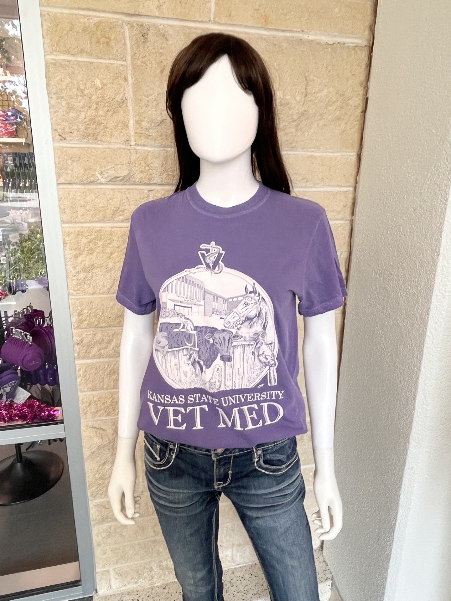A purple short-sleeve graphic T-shirt displayed on a mannequin, paired with blue jeans. The shirt features a light gray graphic of a vet school building and the text 'KANSAS STATE UNIVERSITY VET MED'.