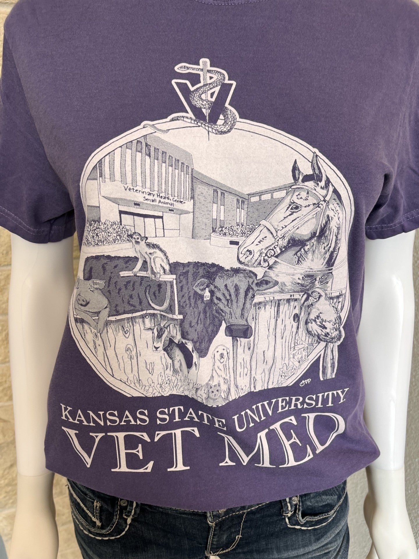 A purple short-sleeve graphic T-shirt displayed on a mannequin torso with dark jeans visible at the bottom. The shirt features a large white circular illustration with a horse, cow, and building, plus the text 'KANSAS STATE UNIVERSITY VET MED' and a snake wrapped around a letter V at the top.