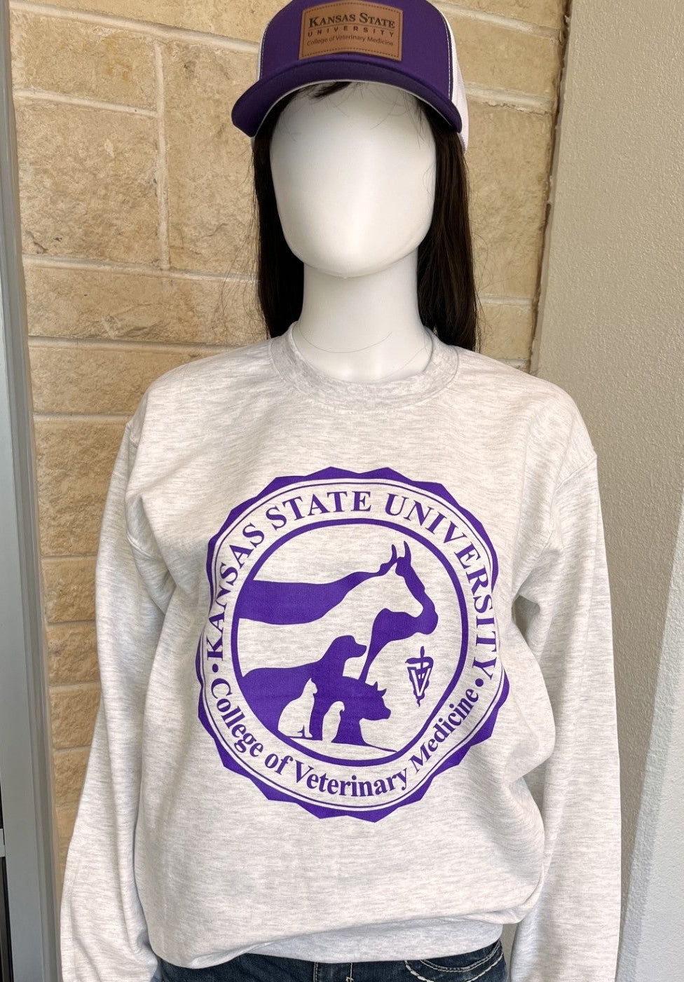 A mannequin wearing a light heather gray sweatshirt with a large purple Kansas State University Veterinary College graphic, paired with dark blue jeans and a purple baseball cap.