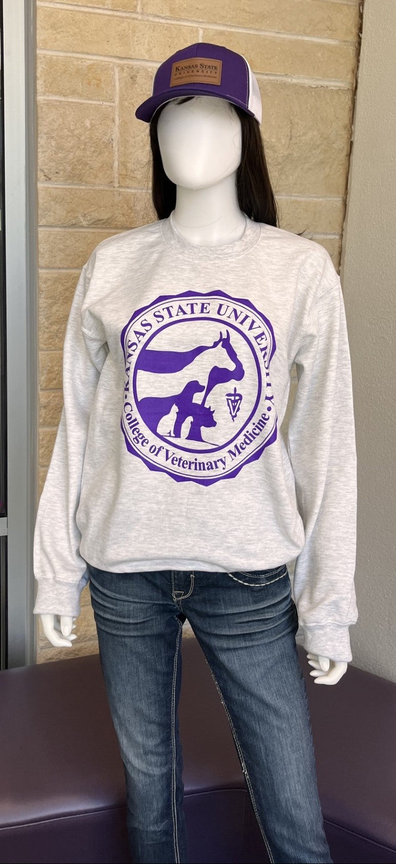 A mannequin wearing a light heather gray sweatshirt with a purple circular Kansas State University Veterinary School graphic, paired with blue jeans and a purple trucker hat, standing indoors near a glass storefront window.