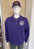 A mannequin wearing a purple quarter-zip pullover with a circular embroidered logo on the left chest, paired with a matching purple trucker hat and dark jeans, displayed in front of a tan stone wall.