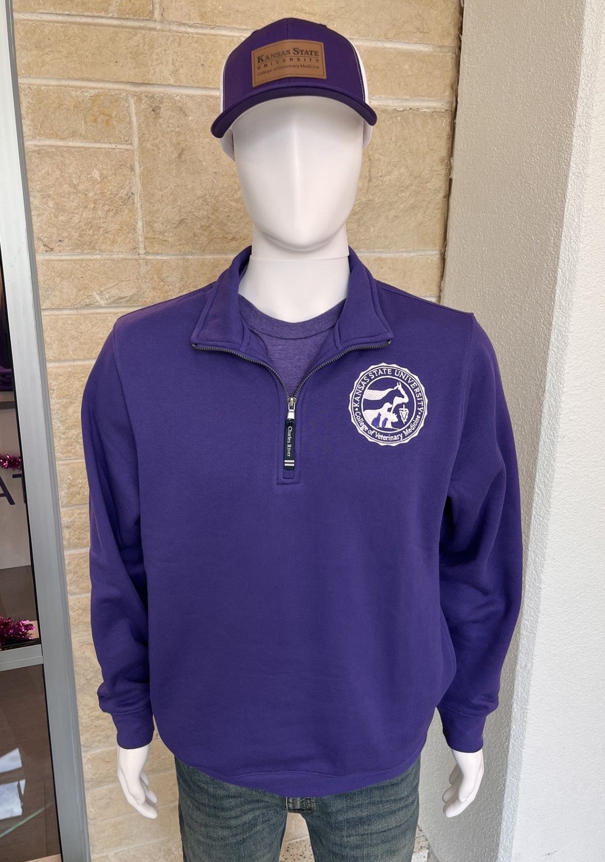 A mannequin wearing a purple quarter-zip pullover with a circular embroidered logo on the left chest, paired with a matching purple trucker hat and dark jeans, displayed in front of a tan stone wall.
