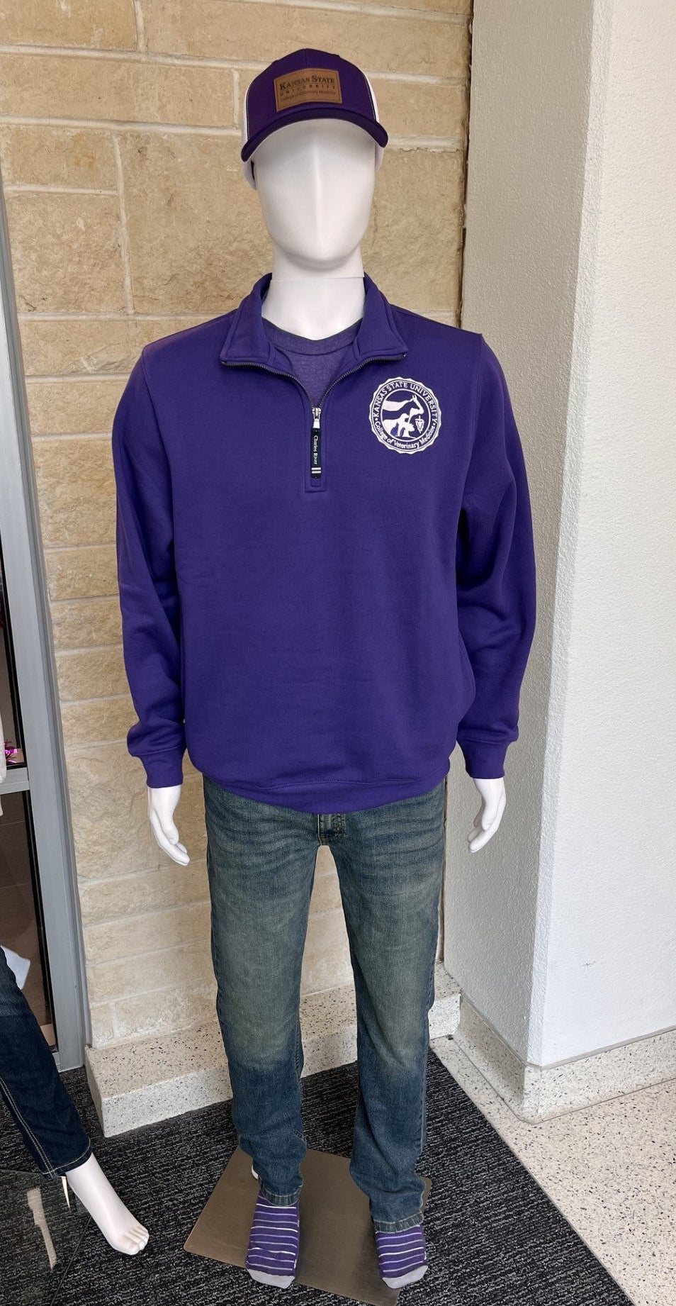 A mannequin wearing a purple quarter-zip pullover with a circular logo on the left chest, blue jeans, and a matching purple cap, displayed in a storefront window area. 