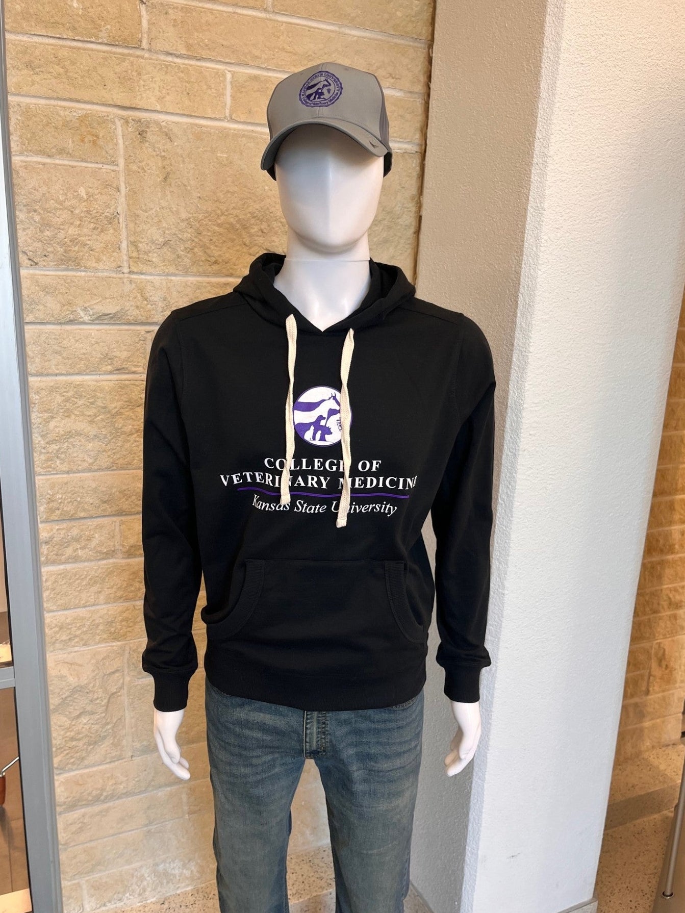 Mannequin wearing a black hoodie with 'College of Veterinary Medicine' text and a CVM seal ballcap, against a beige brick wall.