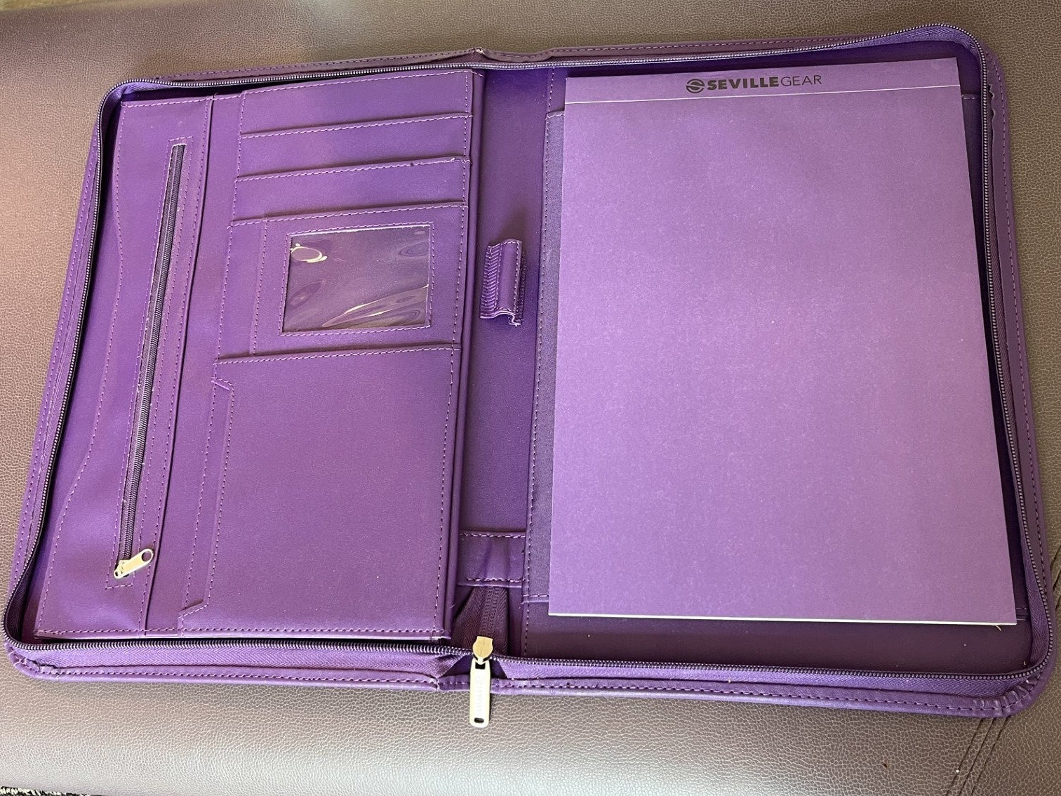 A purple zippered document organizer or tablet sleeve laid open on a beige upholstered chair, showing interior pockets, a clear ID window, and a large front pocket
