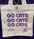 An off-white canvas tote bag with purple text reading 'GO CATS' three times, plus 'Kansas State University' and the Colgate Veterinary Medicine logo at the bottom. The bag has a wooden button closure and is shown hanging against a dark purple background.