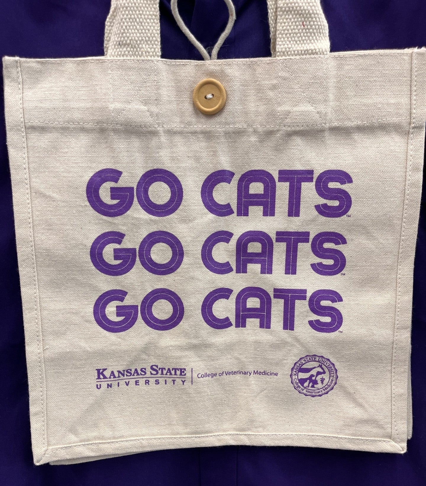 An off-white canvas tote bag with purple text reading 'GO CATS' three times, plus 'Kansas State University' and the Colgate Veterinary Medicine logo at the bottom. The bag has a wooden button closure and is shown hanging against a dark purple background.