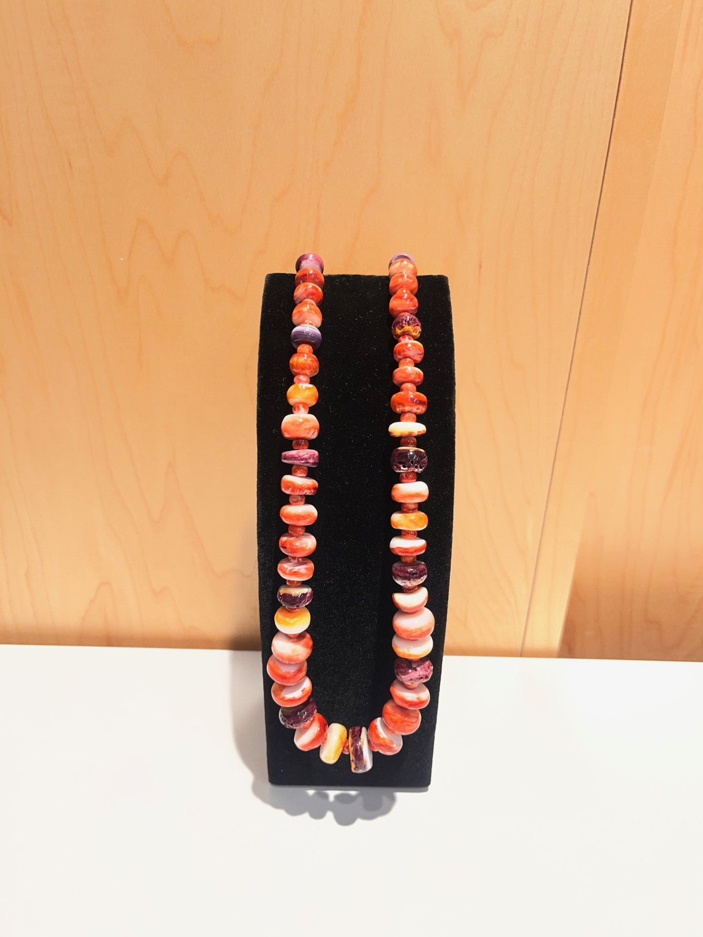 Orange, yellow, red and purple colored oyster beaded necklace on a black stand against a wooden background. 