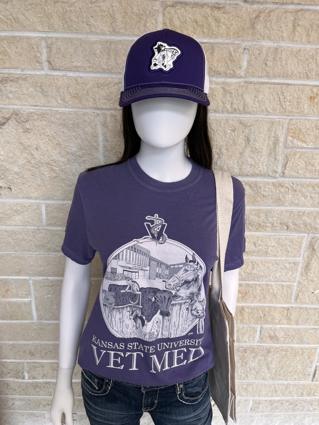 A mannequin wearing a dark purple Kansas State University Vet Med graphic t-shirt, a matching purple baseball cap with a white Lab Coat Willie logo, and dark jeans, standing against a beige stone wall.