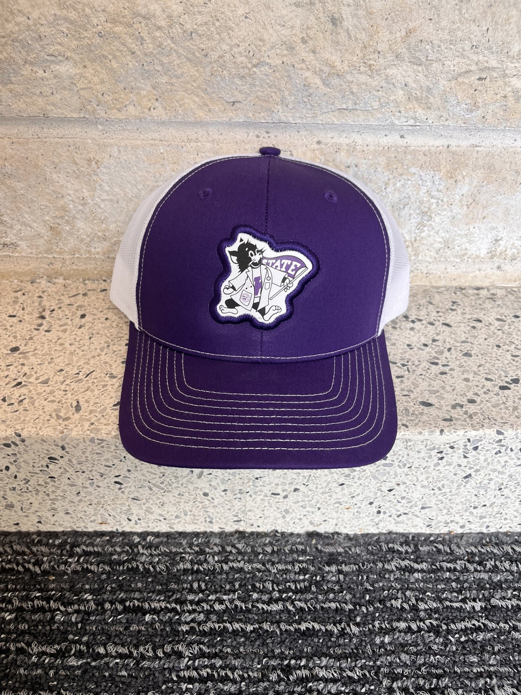 A purple and white trucker hat with a custom embroidered patch on the front, displayed on a speckled stone ledge.