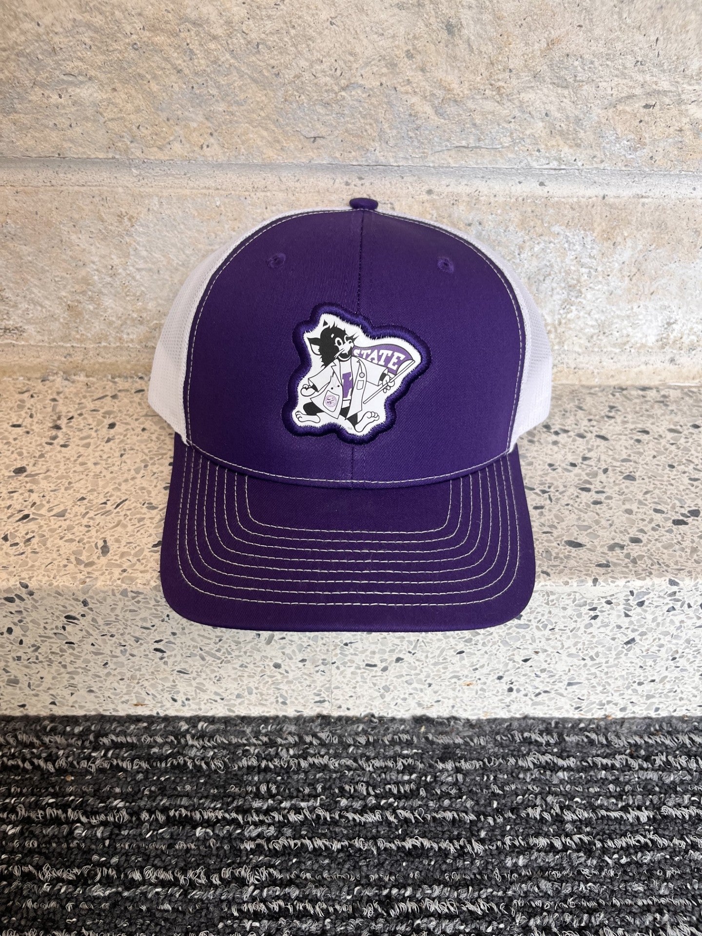 A purple and white trucker hat with a custom embroidered patch on the front, displayed on a speckled stone ledge.