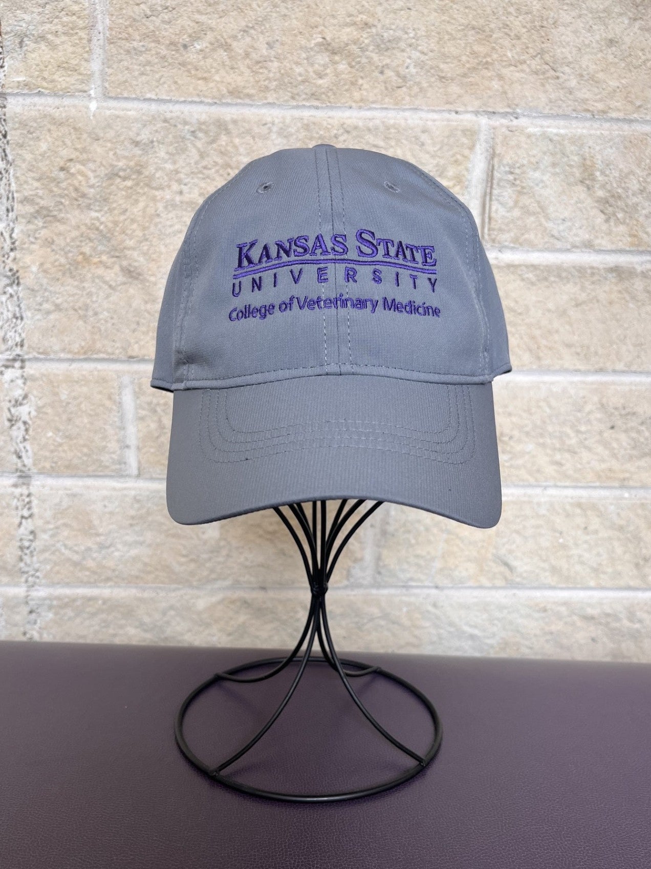 A gray baseball cap with purple embroidered text reading 'Kansas State University College of Veterinary Medicine' displayed on a black wire stand against a beige stone wall background.