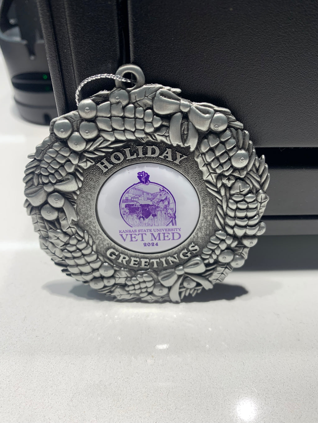 A pewter 2024 Christmas ornament with the text "Holiday Greetings" around a circular center that has an animal image and the text "Kansas State University Vet Med 2024" on a white background. 