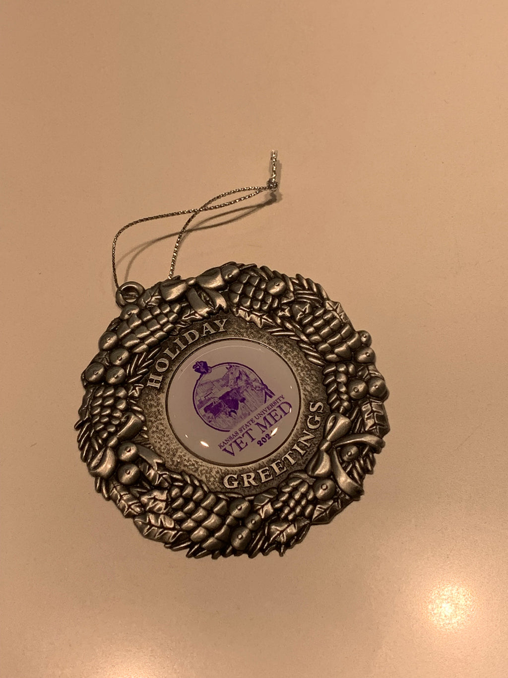 A pewter 2024 Christmas ornament with the text "Holiday Greetings" around a circular center that has an animal image and the text "Kansas State University Vet Med 2024" on a white background. 