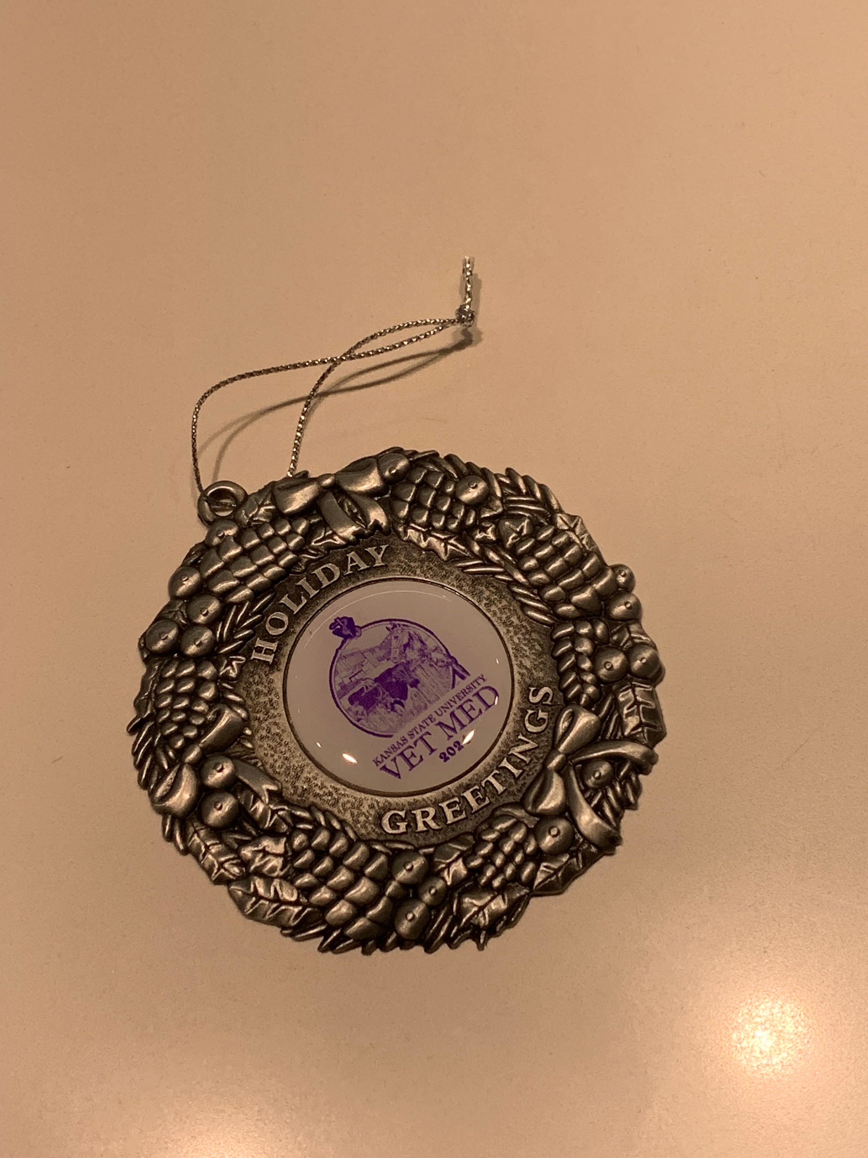 A pewter 2024 Christmas ornament with the text "Holiday Greetings" around a circular center that has an animal image and the text "Kansas State University Vet Med 2024" on a white background. 