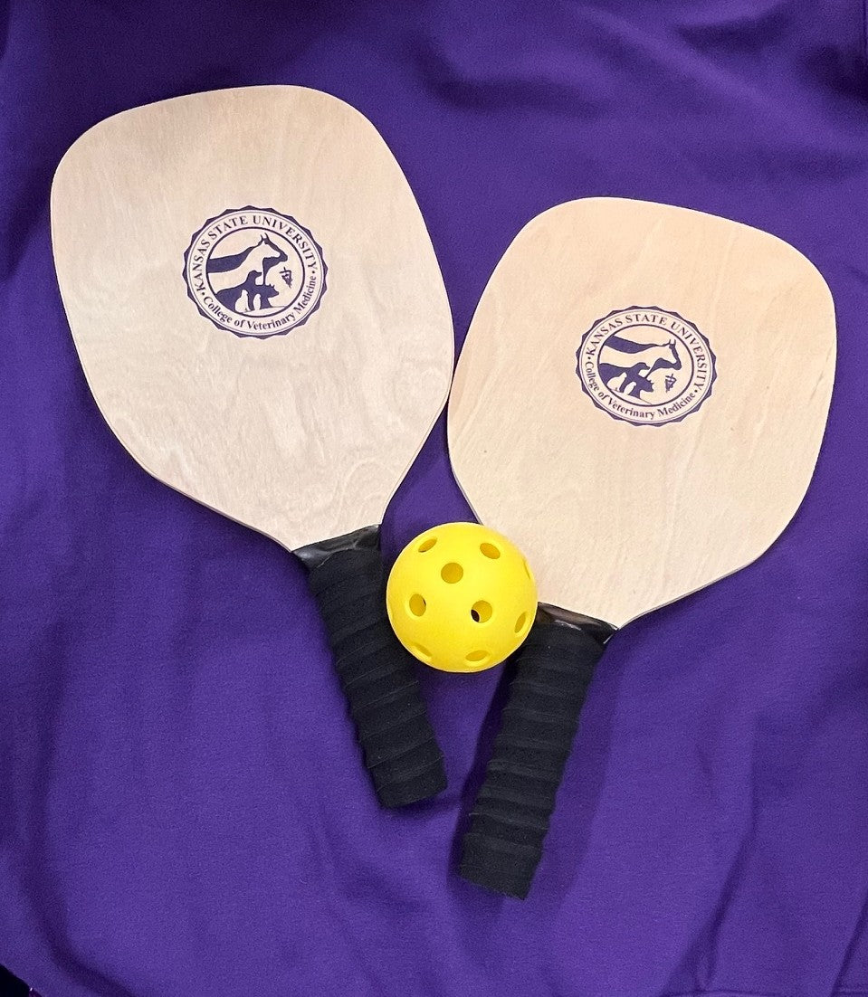 Two wooden pickleball paddles with black handles and a yellow ball on a purple background. The paddles have a dark purple CVM seal in the center. 
