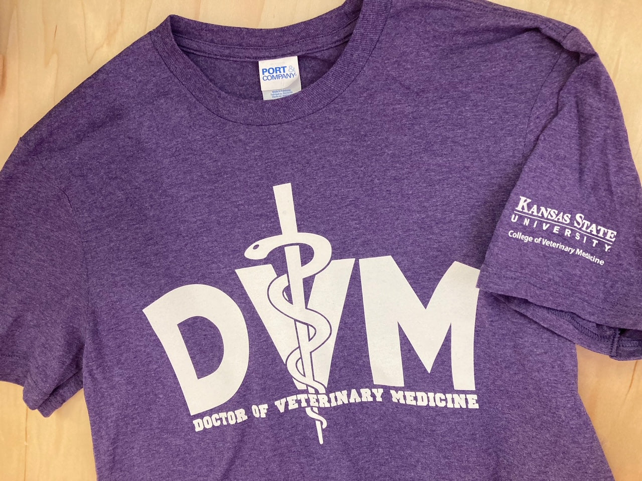 A purple Port & Co. t-shirt with white text that reads "DVM Doctor of Veterinary Medicine" with a white K-State CVM logo on the left sleeve. 
