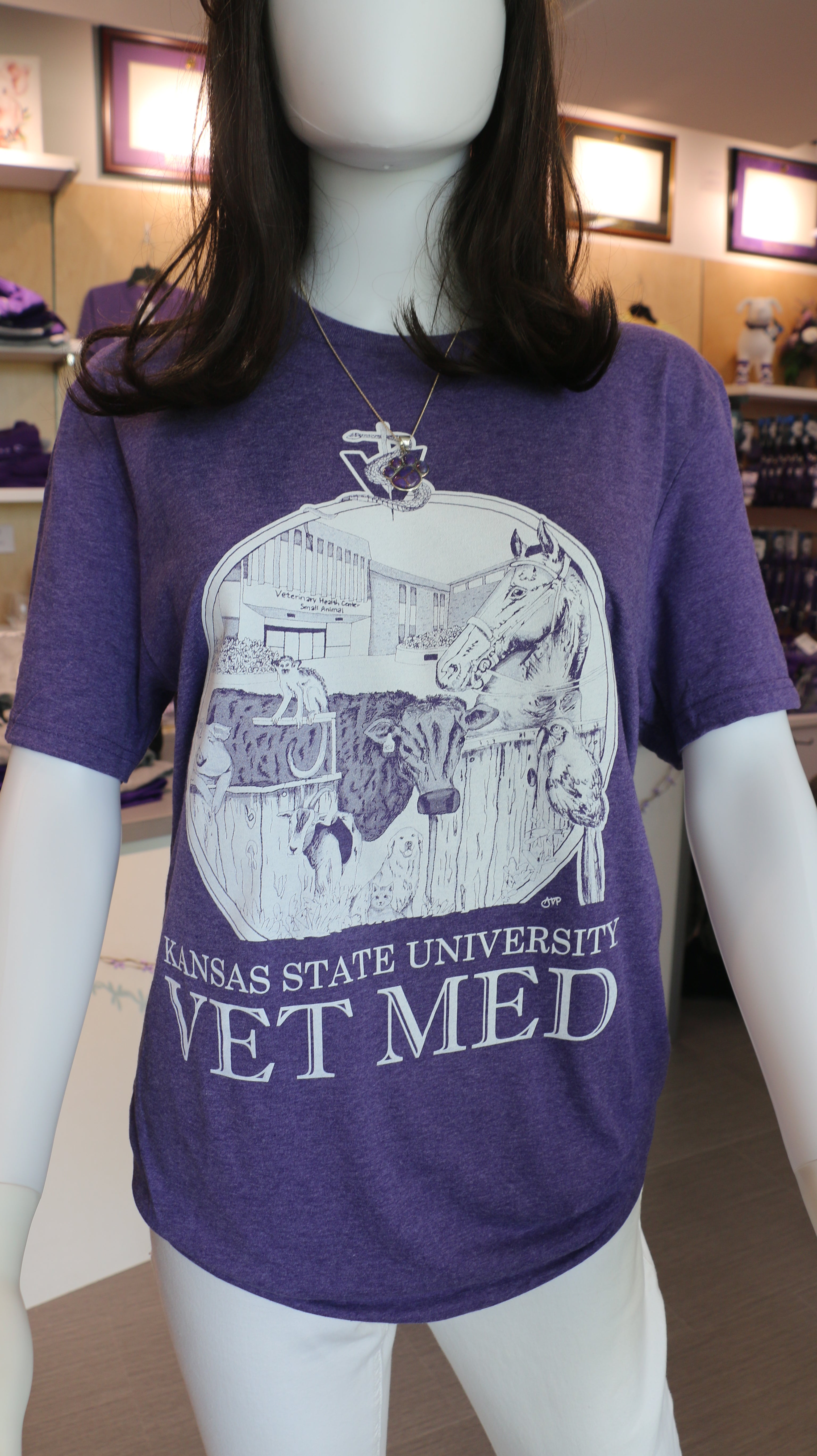 A Port & Company t-shirt featuring a custom design by Ariana Van Pelt, displayed on a mannequin. The shirt is purple with text that reads 'Kansas State University VET MED'.