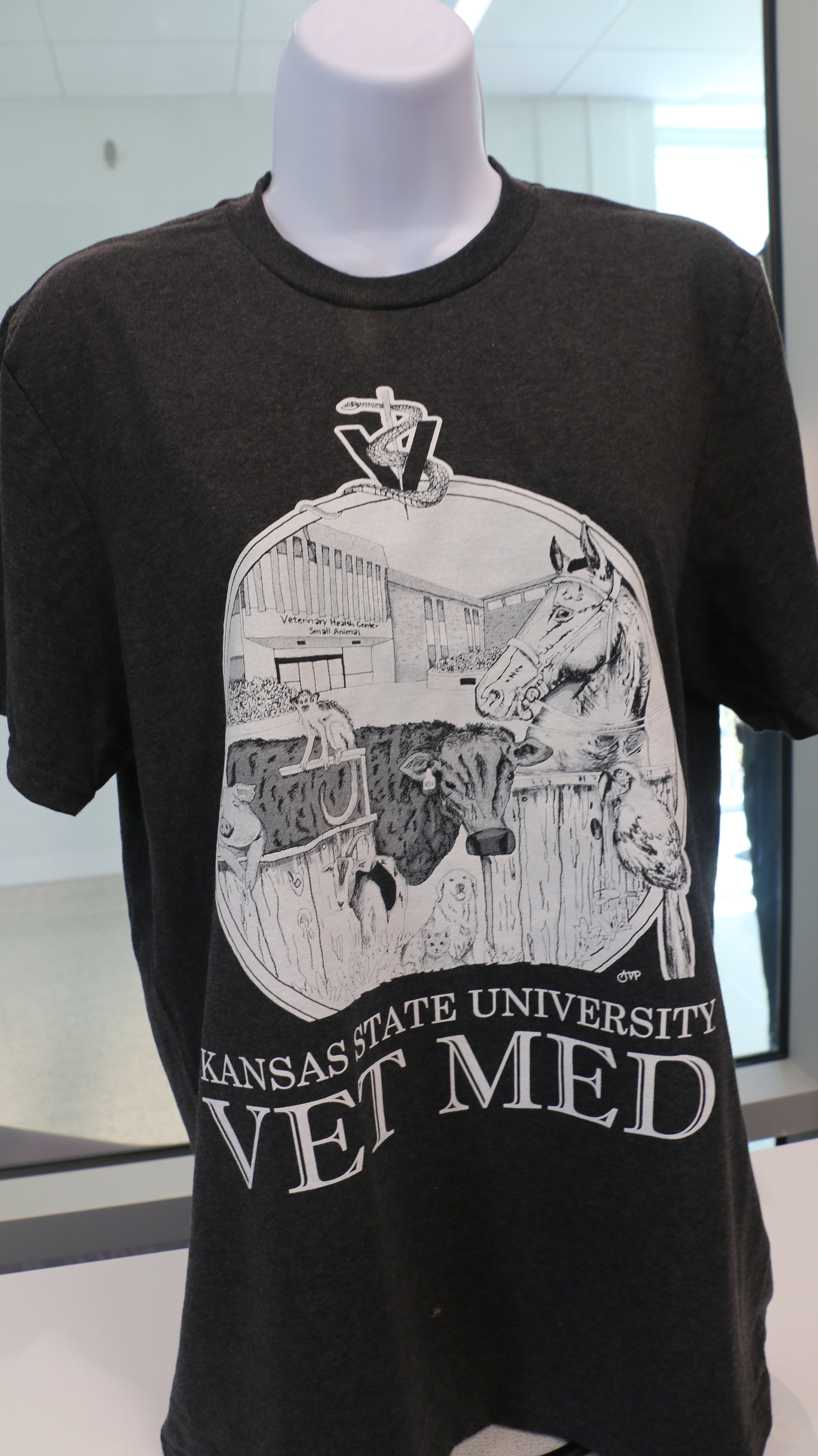 A Port & Company t-shirt featuring a custom design by Ariana Van Pelt, displayed on a mannequin. The shirt is black with text that reads 'Kansas State University VET MED'.
