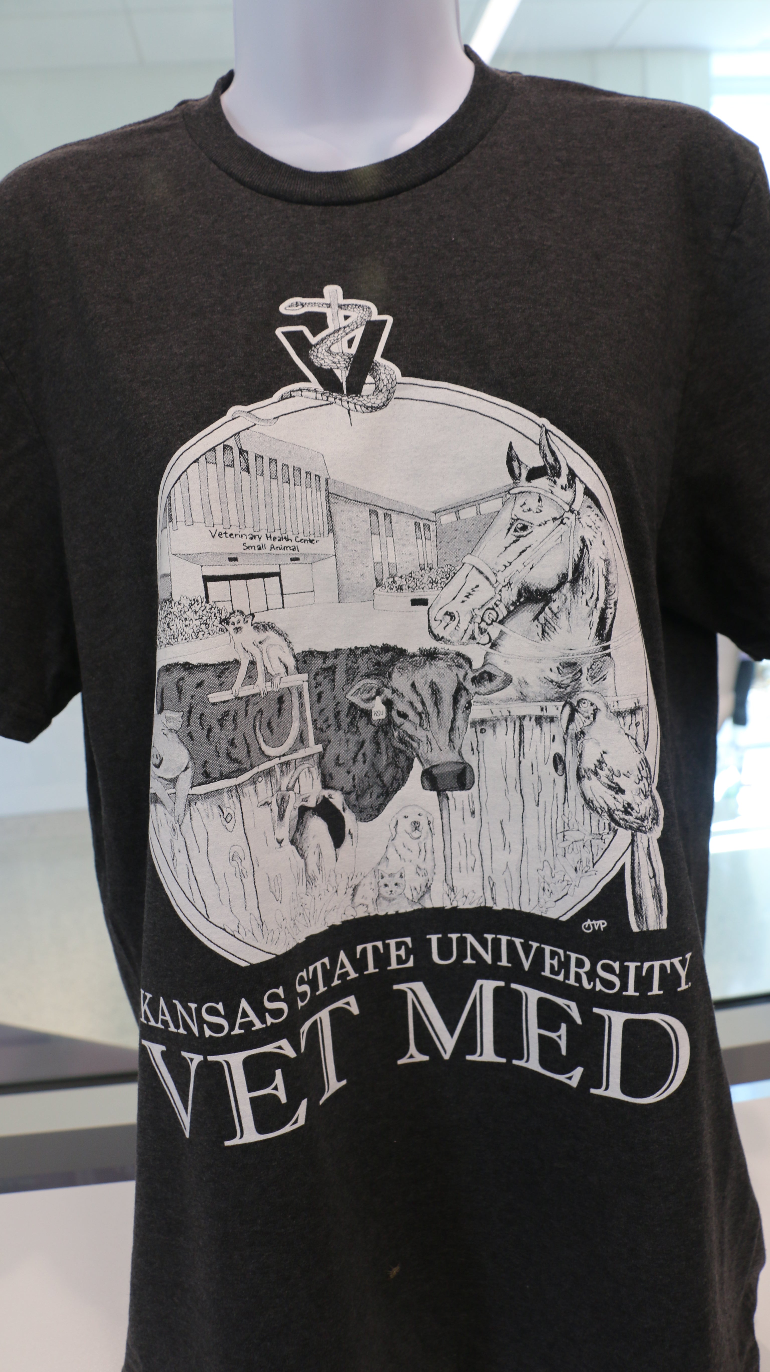 A Port & Company t-shirt featuring a custom design by Ariana Van Pelt, displayed on a mannequin. The shirt is black with text that reads 'Kansas State University VET MED'.