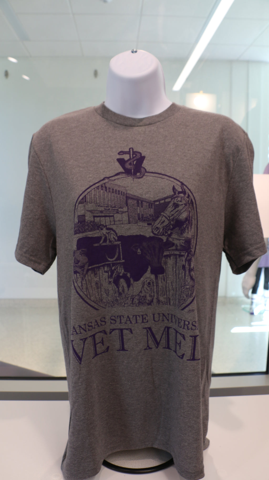 A Port & Company t-shirt featuring a custom design by Ariana Van Pelt, displayed on a mannequin. The shirt is gray with text that reads 'Kansas State University VET MED'.