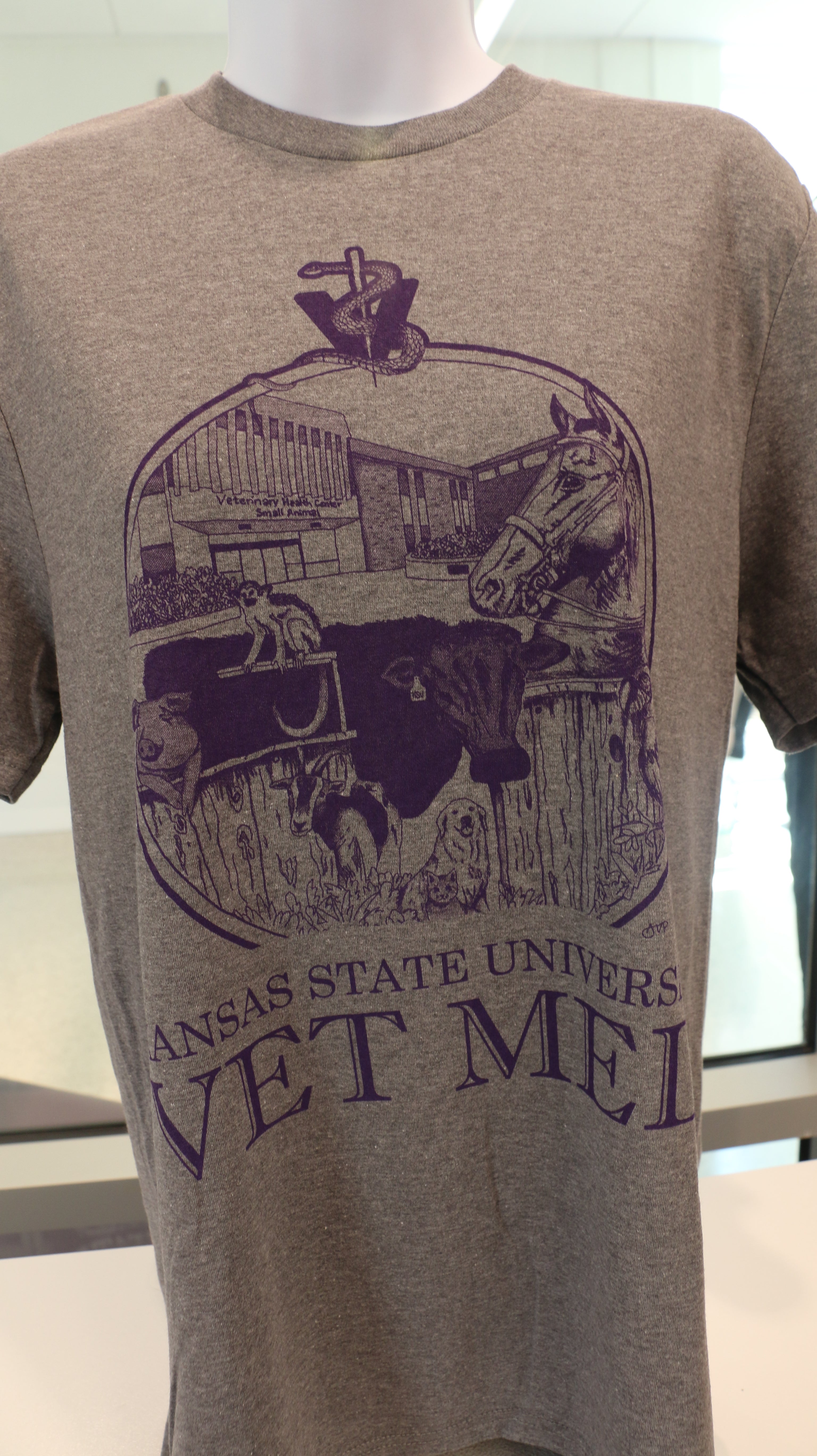 A Port & Company t-shirt featuring a custom design by Ariana Van Pelt, displayed on a mannequin. The shirt is gray with text that reads 'Kansas State University VET MED'.