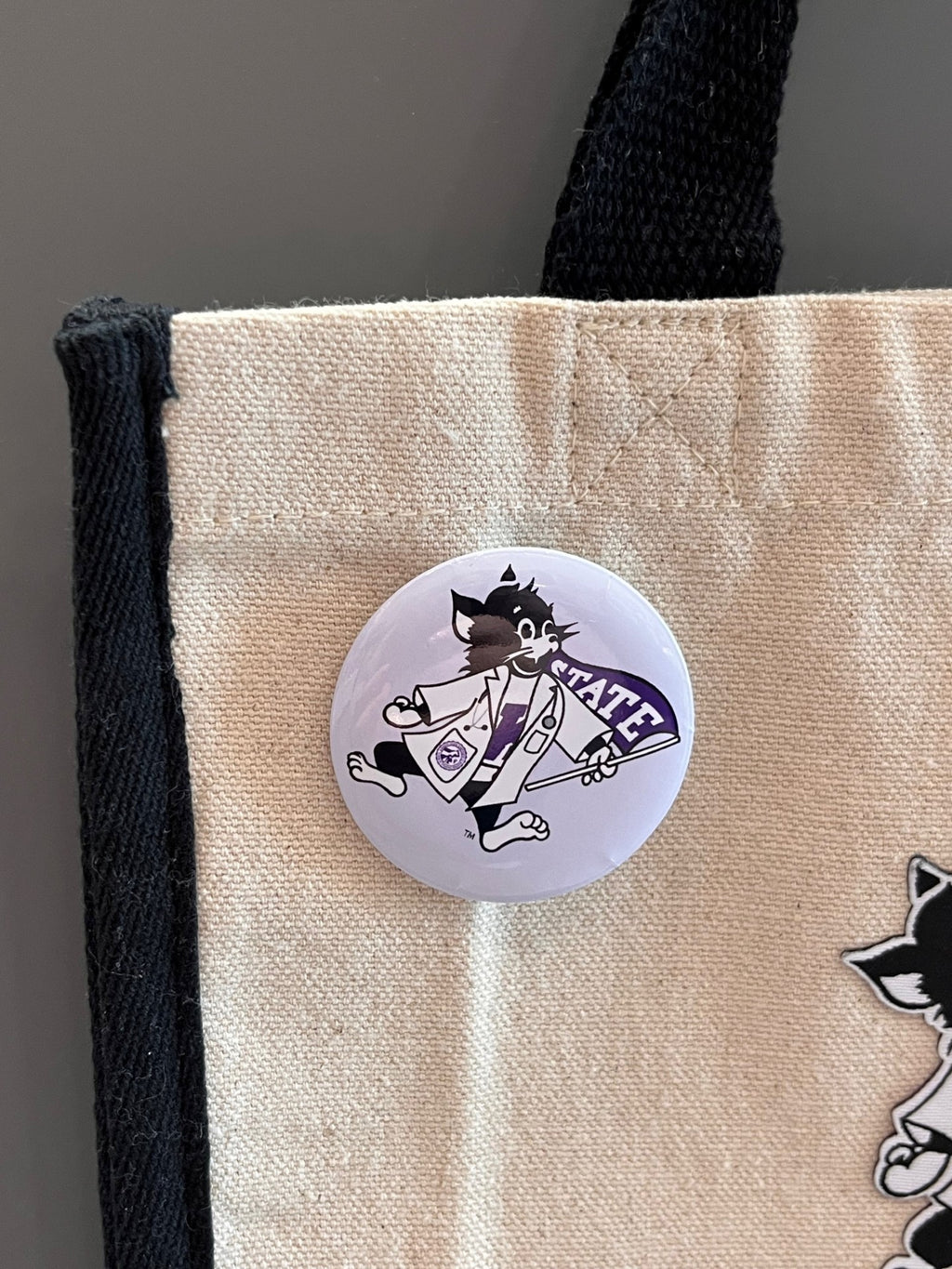 A tote bag with a round button featuring a cartoon Labcoat Willi with a light purple background. 